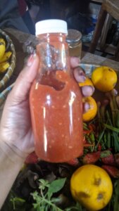 Sebotol sambal luat strobeti yang dipegang. Di latar belakang terdapat buah jeruk asam, cabai, stroberi, dan daun bawang.